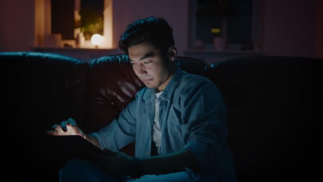 Glücklicher-und-positiver-asiatischer-Kerl-surfen-im-Internet-mit-Tablet-zu-Hause-auf-dem-Sofa.