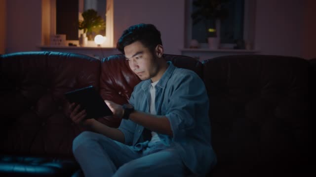 Glücklich-und-Positiv-asiatisch-Kerl-spielen-Spiele-auf-Tablet-zu-Hause-auf-dem-Sofa-in-der-Nacht.