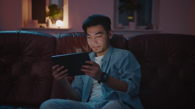 Chico-asiático-feliz-viendo-película-en-línea-en-la-tableta-por-la-noche-sentado-en-el-sofá.
