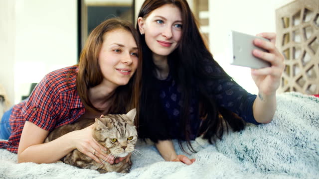 Zwei-glückliche-Frauen-Freunde-im-Bett-liegen-und-dass-Selfie-mit-Katze-und-viel-Spaß-im-Bett-zu-Hause