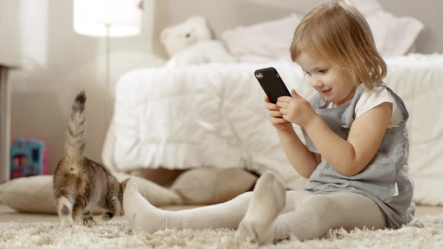 Niedliche-kleine-Girl-Sits-on-the-Floor-mit-Smartphone-und-Shoots-Video-von-Her-Striped-Kitten-Walking-Around.-zeitlupe.