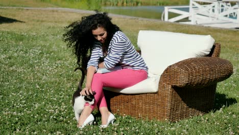 Curly-brunette-relax-in-wicker-armchair-on-green-grass-and-strokes-the-cat