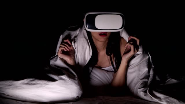 Woman-watching-VR-horror-movie-alone-in-bed.-Asian-woman-lying-in-bed-alone-wearing-VR-head-set,-very-surprise-and-shock-from-scary-movie.-High-tech-Halloween-home-alone-concept.