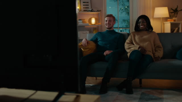 Happy-Diverse-Young-Couple-Watching-Comedy-on-TV-while-Sitting-on-a-Couch,-they-Laugh-and-Enjoy-Show.-Handsome-Caucasian-Boy-and-Black-Girl-in-Love-Spending-Time-Together-in-the-Cozy-Apartment.