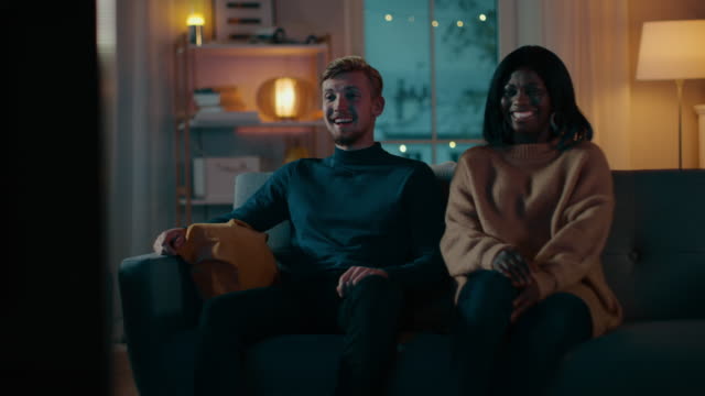 Happy-Diverse-Young-Couple-Watching-Comedy-on-TV-while-Sitting-on-a-Couch,-they-Laugh-and-Enjoy-Show.-Handsome-Caucasian-Boy-and-Black-Girl-in-Love-Spending-Time-Together-in-the-Cozy-Apartment.