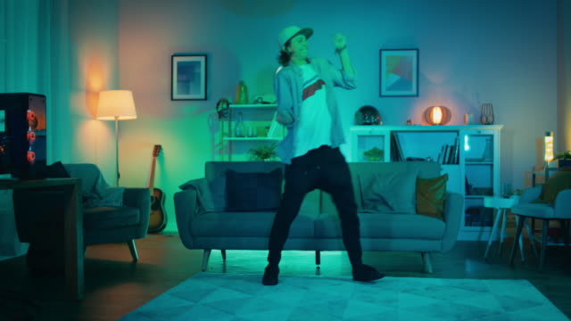 Handsome-Excited-Hip-Young-Man-is-Dancing-in-the-Living-Room-while-TV-Plays-in-the-Background.-He-is-Energetically-Moving-while-Screen-Adds-Reflections-on-Him.-Cozy-Room-is-Lit-with-Warm-Neon-Light.