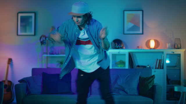 Handsome-Excited-Hip-Young-Man-is-Dancing-in-the-Living-Room-while-TV-Plays-in-the-Background.-He-is-Energetically-Moving-while-Screen-Adds-Reflections-on-Him.-Cozy-Room-is-Lit-with-Warm-Neon-Light.