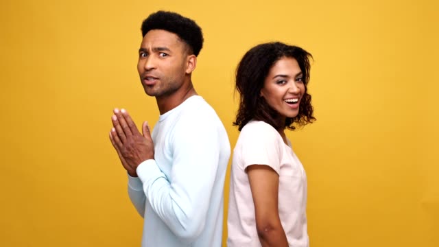 Young-african-couple-standing-back-to-back,-man-showing-disagreement-and-woman-showing-approval-gesture-isolated-over-yellow-background