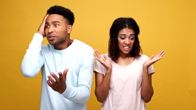 Frustrated-young-afro-american-man-having-a-disagreement-with-his-guilty-girlfriend-isolated-over-yellow-background