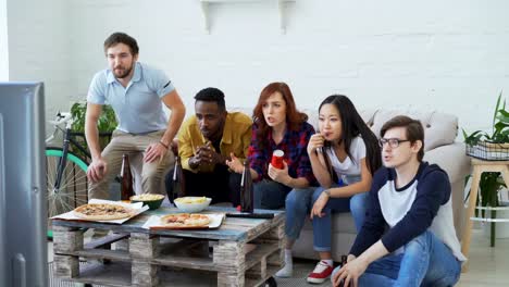 Group-of-young-friends-watching-olympic-games-match-on-TV-together-eating-snacks-and-drinking-beer.-Some-of-them-happy-with-their-team-winning-but-others-disappointed