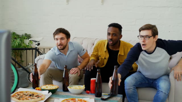 Gruppe-von-jungen-männlichen-Freunden-Sportübertragungen-im-Fernsehen-zusammen-passen-beim-trinken-Bier-und-Snacks-zu-Hause-essen