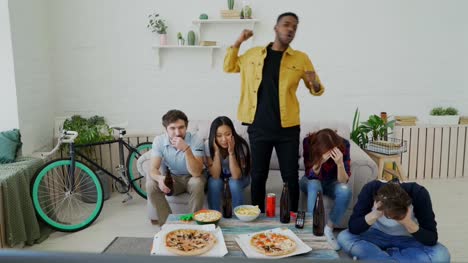 Group-of-young-friends-watching-olympic-games-match-on-TV-together-eating-snacks-and-drinking-beer.-African-man-is-happy-about-his-team-winning-but-others-are-disappointed