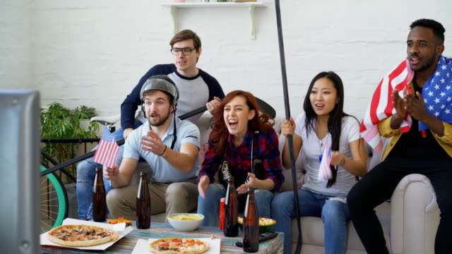 Multi-ethnic-group-of-friends-sports-fans-with-USA-national-flags-watching-winter-olympic-games-on-TV-together-cheering-up-favourite-team-at-home