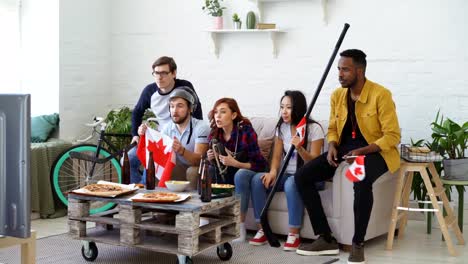 Multi-ethnische-Gruppe-von-Freunden-Sport-Fans-mit-kanadischen-Nationalflaggen-beobachten-Hockey-Meisterschaft-im-Fernsehen-zusammen-ihre-Lieblingsmannschaft-zu-Hause-Aufmunterung