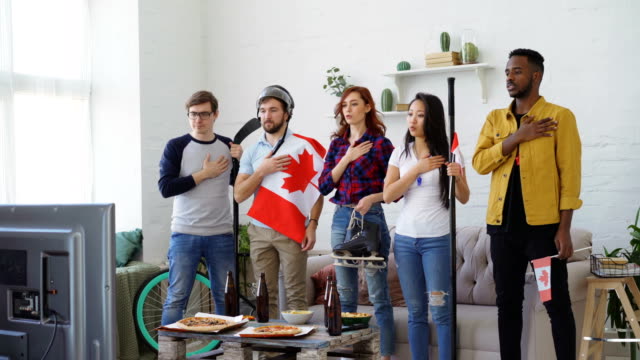 Multi-ethnic-group-of-friends-listening-and-singing-national-Canadian-anthem-before-watching-sports-championship-on-TV-together-at-home