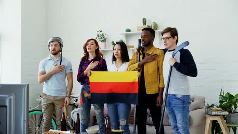 Multi-ethnic-group-of-friends-listening-and-singing-German-national-anthem-before-watching-sports-championship-on-TV-together-at-home