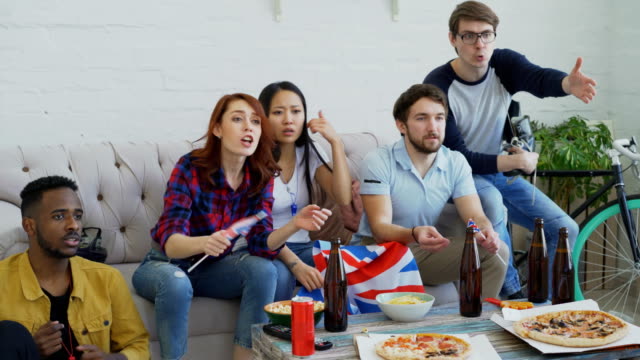 Group-of-young-friends-sports-fans-with-British-national-flags-watching-sport-championship-on-TV-together-and-happy-about-winning-favourite-team-at-home
