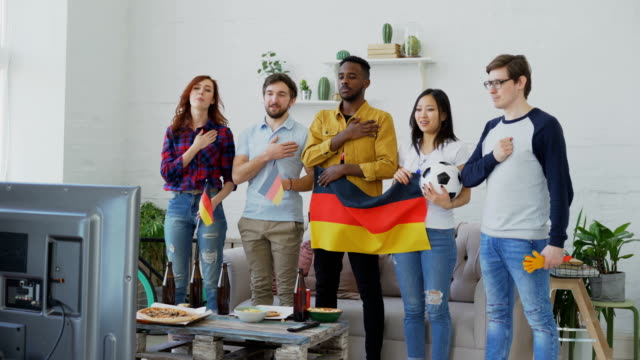 Multi-ethnischen-Gruppe-von-Freunden-hören-und-singen-der-deutschen-Nationalhymne-vor-gerade-Sport-Meisterschaft-im-Fernsehen-zusammen-zu-Hause