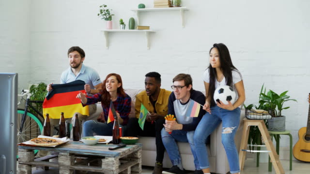Amigos-jóvenes-viendo-Juegos-Olímpicos-partido-en-TV-juntos-en-casa.-Algunos-de-ellos-contento-con-el-equipo-alemán-en-ganar-pero-hombre-brasileño-decepcionó