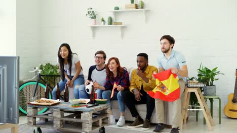 Multi-ethnic-group-of-friends-sports-fans-with-Spanish-flags-watching-football-championship-on-TV-together-at-home-and-cheering-up-favourite-team