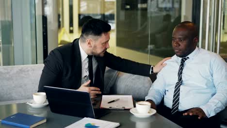 Angry-bussinessman-in-suit-scolding-his-african-american-employee-and-grabs-his-tie-pointing-on-fail-in-contract-at-modern-cafe.-Angry-boss-striking-table-with-his-hand-nervously