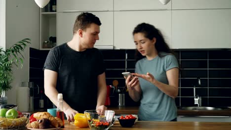 young-handsome-caucasian-man-standing-at-table-cutting-vegetables-and-trying-piece-of-yellow-pepper,-his-girlfriend-comes-furious-holding-mobile-phone,-man-laughing