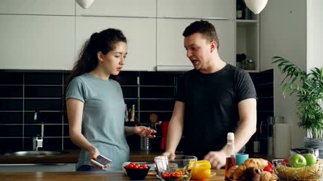young-curly-woman-showing-something-unpleasant-in-husband's-phone-while-he-is-cooking-,-they-are-shouting-and-quarrelling,-man-is-angry-and-irritated