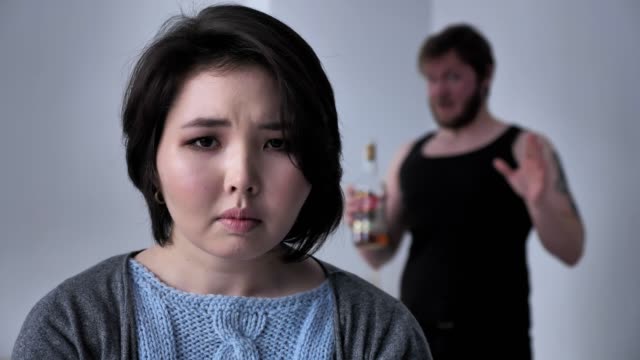 Portrait-of-a-sad-depressed-Asian-girl,-drunk-husband-in-the-background-swears,-alcohol,-conflict,-looks-at-the-camera.-50-fps