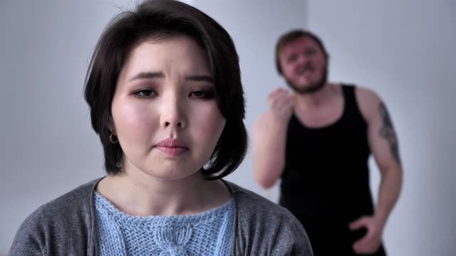 Portrait-of-a-sad-depressed-Asian-girl,-drunk-husband-in-the-background-is-grieving,-alcohol,-violence,-looks-at-the-camera.-60-fps
