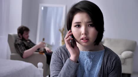 Young-sad-Asian-girl-anxiously-dials-a-number,-calls-by-smartphone,-portrait,-looks-at-camera,-drunk-husband-in-the-background-50-fps