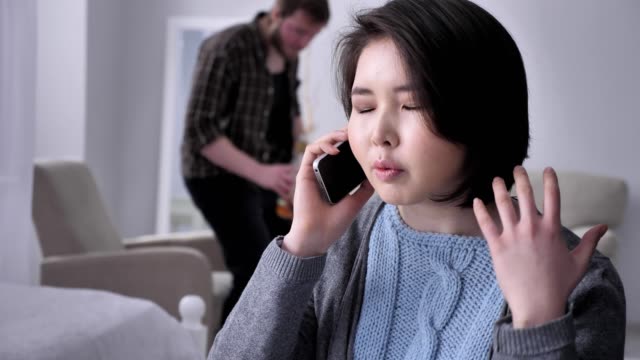 Young-sad-Asian-girl-anxiously-talking-on-the-phone-using-smartphone-smartphone,-portrait,-drunk-husband-in-background-50-fps