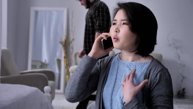Young-sad-Asian-girl-speaks-aggressively-on-the-phone-using-smartphone,-portrait,-drunk-husband-in-background-50-fps