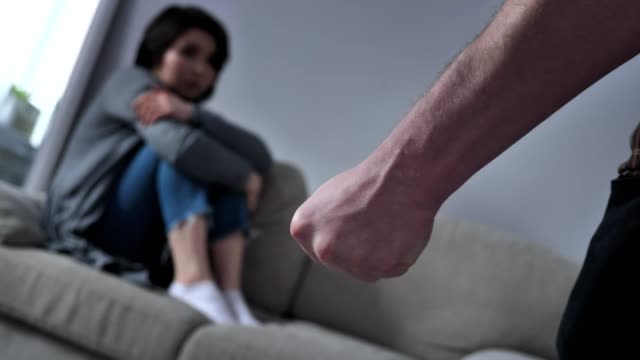 Concept-of-domestic-violence-in-family,-male-fist-afraid-Asian-woman-sitting-on-the-couch-in-the-background-50-fps