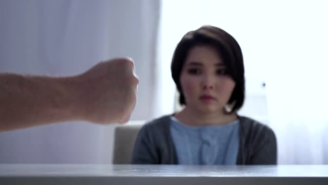 Konzept-von-häuslicher-Gewalt-in-der-Familie,-schlägt-Faust-auf-Tisch,-ängstlich-asiatische-Frau-sitzt-am-Tisch-im-Hintergrund-50-fps