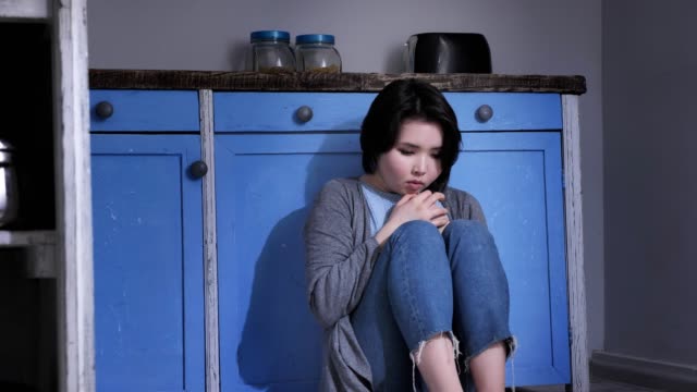 Sad-lonely-young-Asian-girl-sitting-on-the-floor-in-kitchen,-holding-her-knees-with-arms,-domestic-violence-concept-50-fps