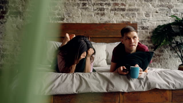 Young-man-is-watching-TV-with-his-girlfriend-lying-on-bed-and-holding-remote,-they-are-holding-pillows-and-mug.-Television,-family-life-and-entertainment-concept.
