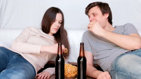 Couple-young-man-and-woman-lie-on-the-floor-at-home-eating-chips-and-drinking-beer-and-talking.