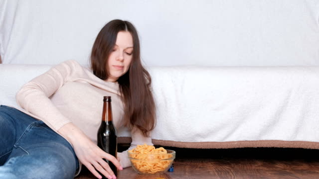 Young-beautiful-woman-drinking-a-beer-and-eating-chips-laying-on-the-floor-near-the-couch.