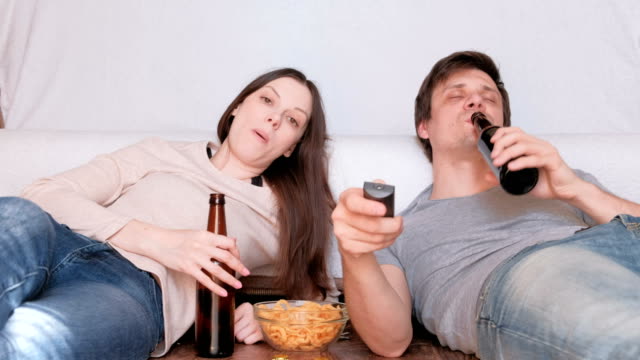 Couple-of-young-man-and-woman-eating-chips-drinking-beer-and-watching-TV,-switching-channel.-Talking-and-smiling