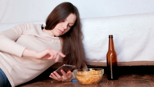 Sad-young-beautiful-brunette-woman-typing-a-message-on-the-mobile-phone,-drinks-beer-and-eats-chips.