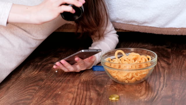 Young-woman-typing-a-message-on-the-mobile-phone,-eating-chips-and-drinking-beer.-Closeup-hands.