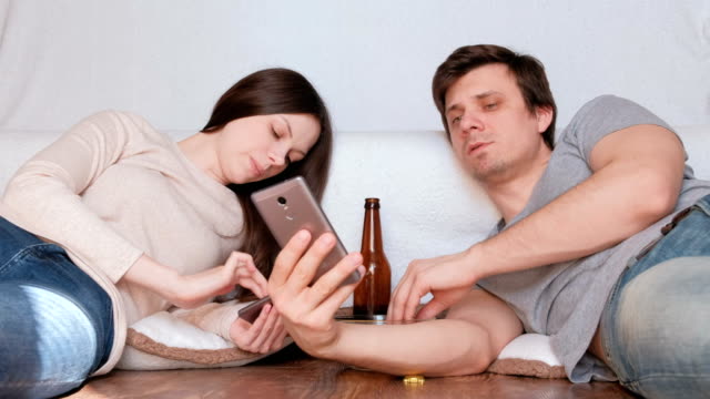 Pareja-hombre-y-mujer-navegar-por-internet-en-sus-teléfonos-móviles,-bebiendo-cerveza-y-comiendo-fritas-tendido-en-el-piso.
