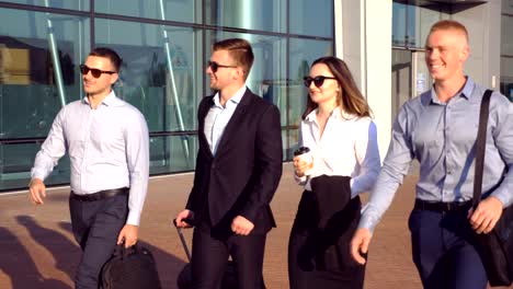 Group-of-young-colleagues-walking-at-city-street-during-sunny-day.-Close-up-of-business-partners-being-on-his-way-to-business-trip.-Coworkers-going-near-airport-terminal-and-talking.-Slow-motion