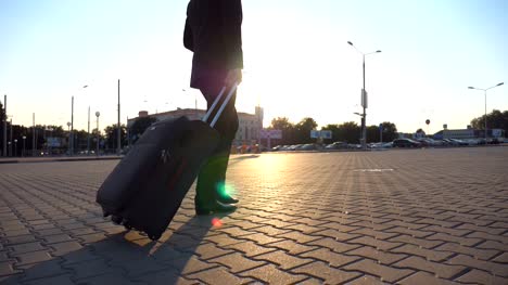 Profile-of-successful-businessman-in-a-black-suit-walking-with-his-luggage-on-urban-street-at-sunset.-Confident-young-man-going-to-airport-terminal-and-pulling-suitcase-on-wheels.-Close-up-Side-view