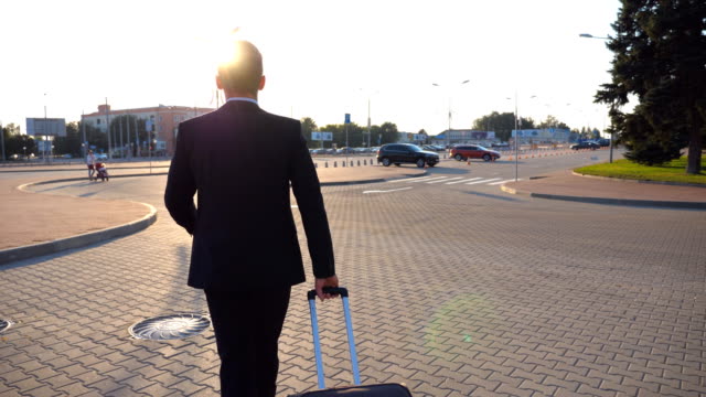 Rear-view-of-unrecognizable-young-businessman-walking-to-airport-terminal-and-pulling-suitcase-on-wheels-at-sunset.-Successful-male-business-person-going-with-his-luggage-on-city-street.-Close-up