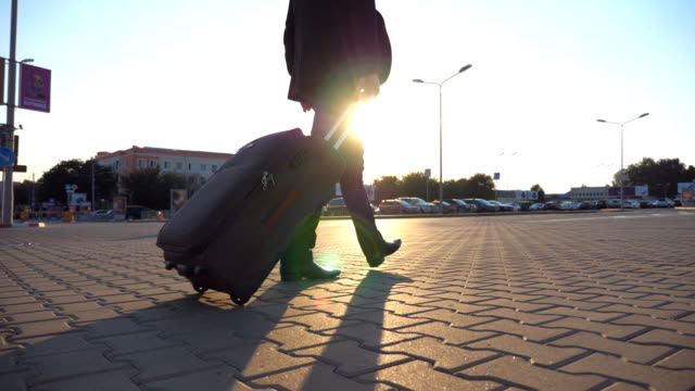 Crane-shot-of-unrecognizable-man-walking-on-city-street-and-pulling-suitcase-on-wheels-at-sunset.-Young-businessman-looking-at-his-watch-and-going-with-luggage-to-airport-terminal.-Close-up-Rear-view