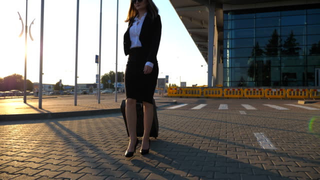 Young-woman-walking-near-airport-parking-with-her-luggage-at-sunset-time.-Female-legs-in-high-heels-stepping-at-sidewalk.-Business-woman-going-with-her-suitcase-on-urban-street.-Slow-motion-Close-up