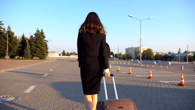 Business-woman-with-her-suitcase-going-from-airport-to-taxi-parking.-Lady-walking-with-her-luggage-along-city-street.-Female-legs-in-high-heels-stepping-at-sidewalk.-Rear-back-view-Slow-motion