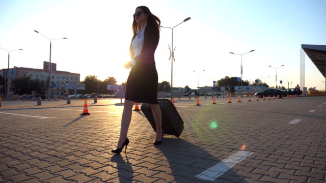 Woman-in-high-heeled-footwear-walking-near-airport-parking-with-her-luggage-at-sunset-time.-Businesswoman-going-with-her-suitcase-along-city-street.-Sun-flare-at-background.-Slow-motion-Close-up
