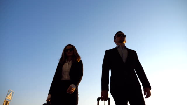 Business-man-and-woman-going-to-the-airport-with-their-luggage.-Young-businessman-carrying-suitcase-on-wheels-and-walking-with-his-female-colleague-to-terminal-hall.-Trip-or-travel-concept.-Low-angle-view-Slow-motion